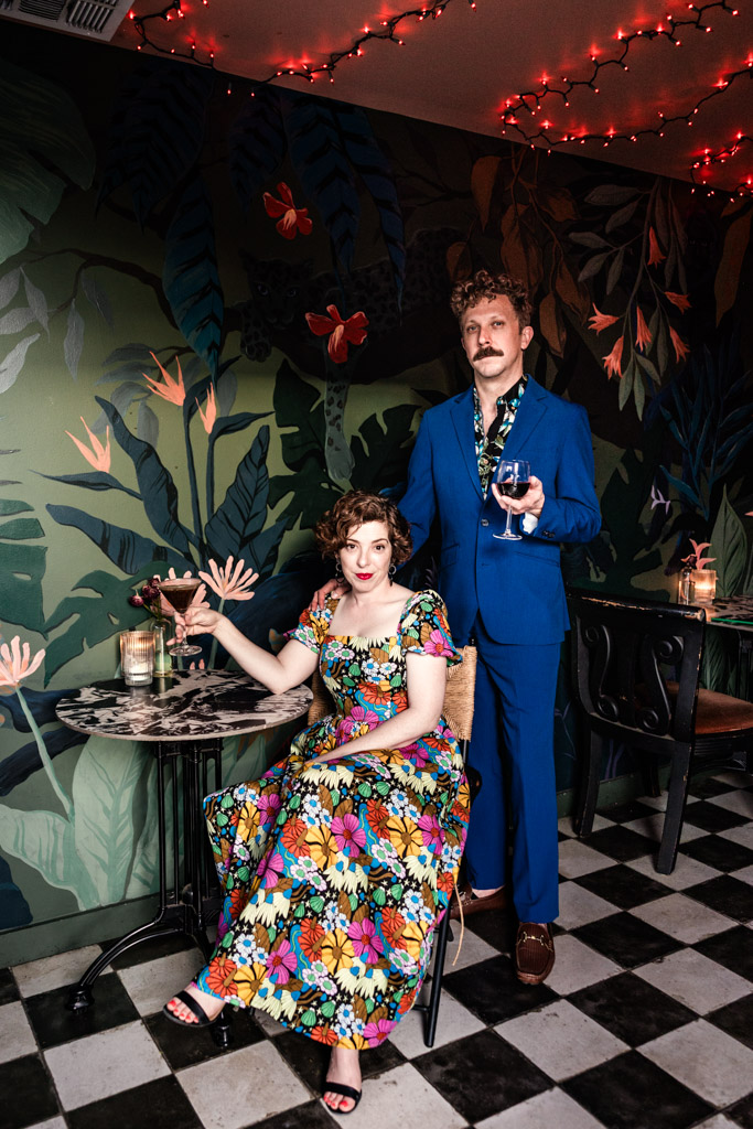 Portrait of woman in a floral dress sitting at a table with a man in a blue suit standing beside her, both in a colorful, decorated room at Justine's wine bar in Los Angeles