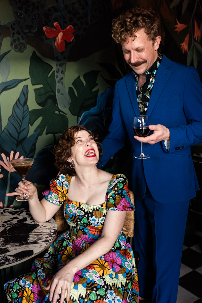 A smiling woman in a floral dress holds a drink, while a man in a blue suit stands beside her with a wine glass at Justine's in Los Angeles