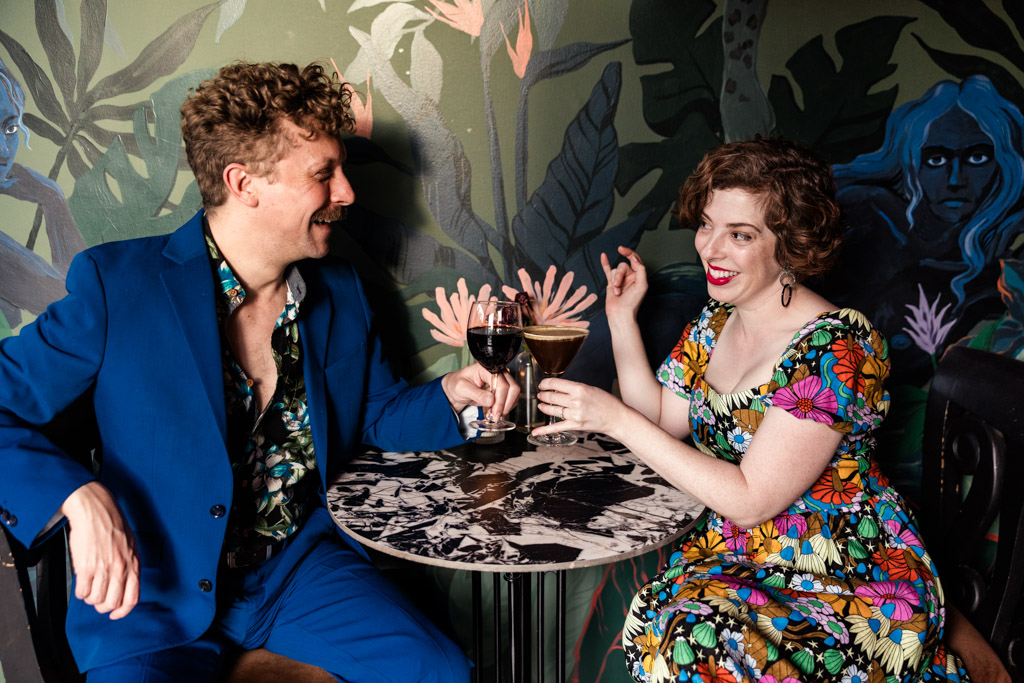 A man and woman sit at a small table, smiling and toasting drinks in Justine's wine bar