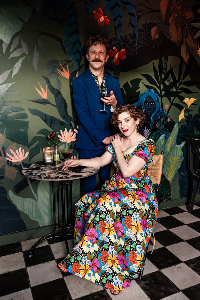 Portrait of man in a blue suit and a woman in a floral dress sitting at a small table in front of a vibrant mural inside Justine's