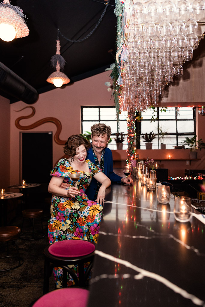 Two people smile and sit at the bar inside Justine's with colorful decor, candles, and glasses hanging above
