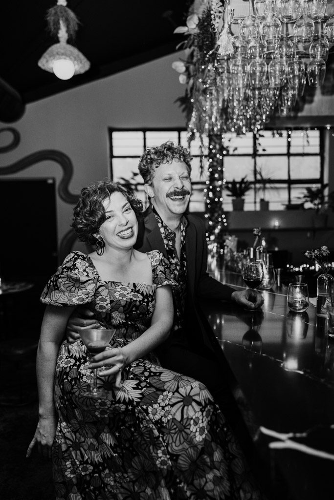 Black and white photo of happy engaged couple in floral outfits sitting at a barInside Justine's in Los Angeles