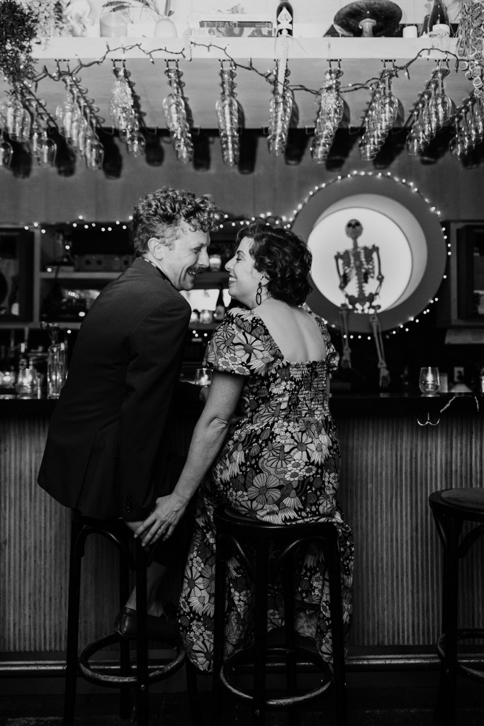 Black and white photo of engaged couple sitting close together at a bar inside Justine's, smiling at each other, with a skeleton decoration in the background