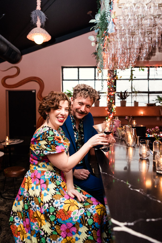 A smiling couple in colorful outfits sit at a bar holding drinks, surrounded by festive lights and decor