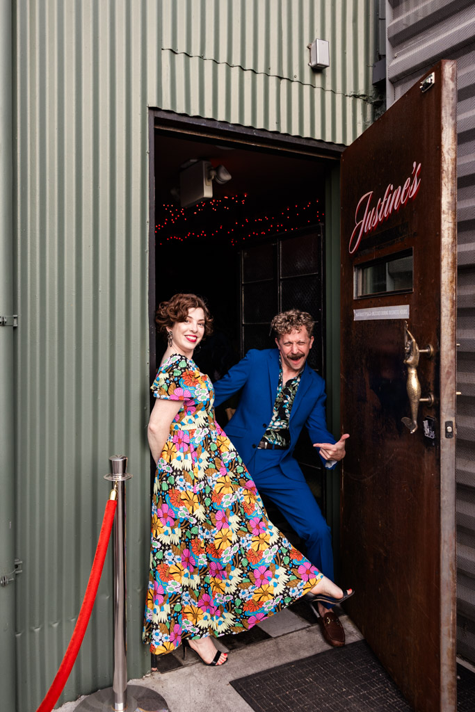 A smiling woman in a floral dress and a man in a blue suit pose playfully at the entrance of Justine's wine bar in Los Angeles