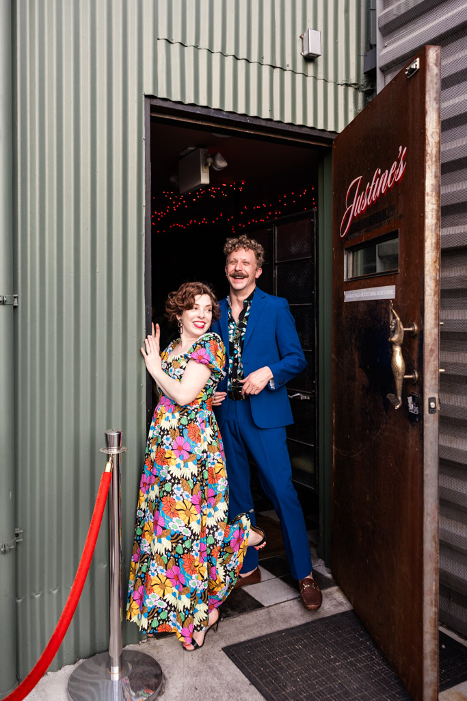 A smiling couple in colorful outfits stands at the entrance of Justine’s, near a red rope during Los Angeles engagement session