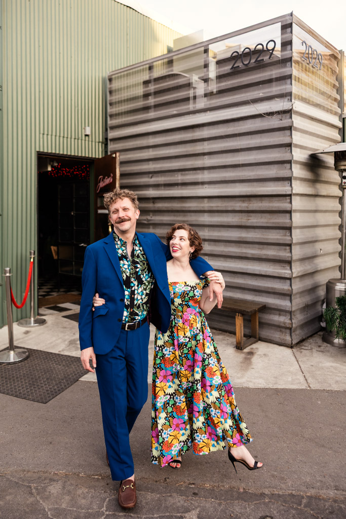 A smiling couple in colorful outfits walks arm-in-arm outside Justine's during their Los Angeles engagement session