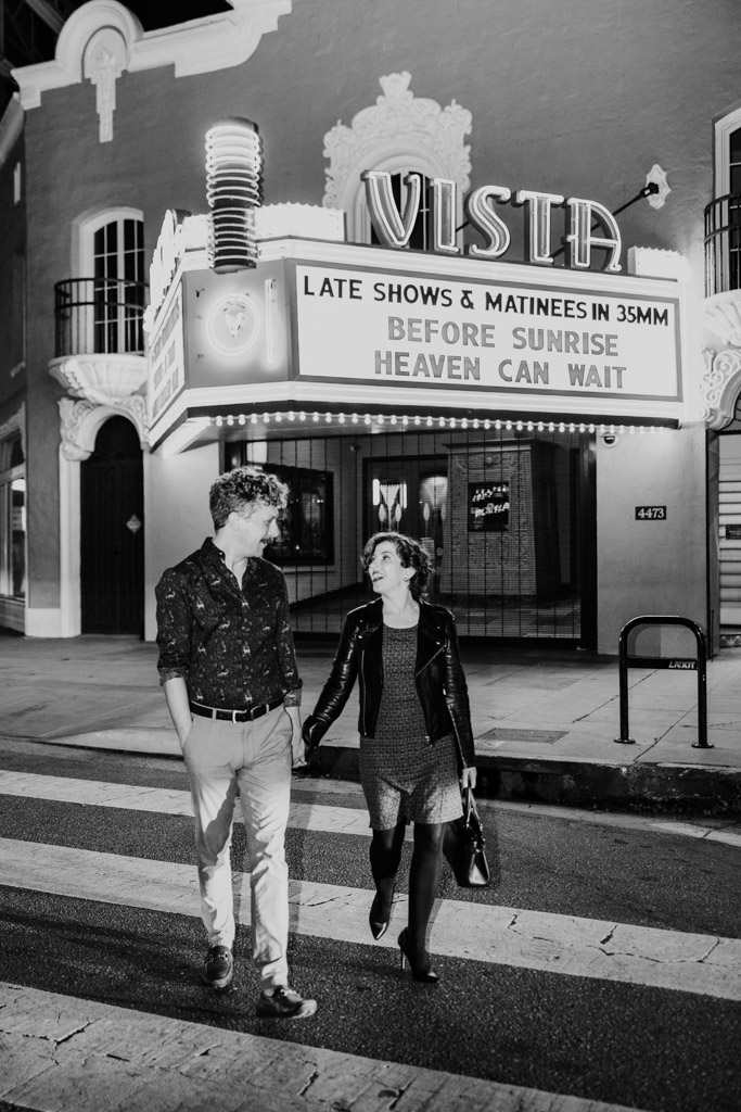 Black and white photo of engaged couple holding hands walking past the Vista Theatre at night under a glowing marquee