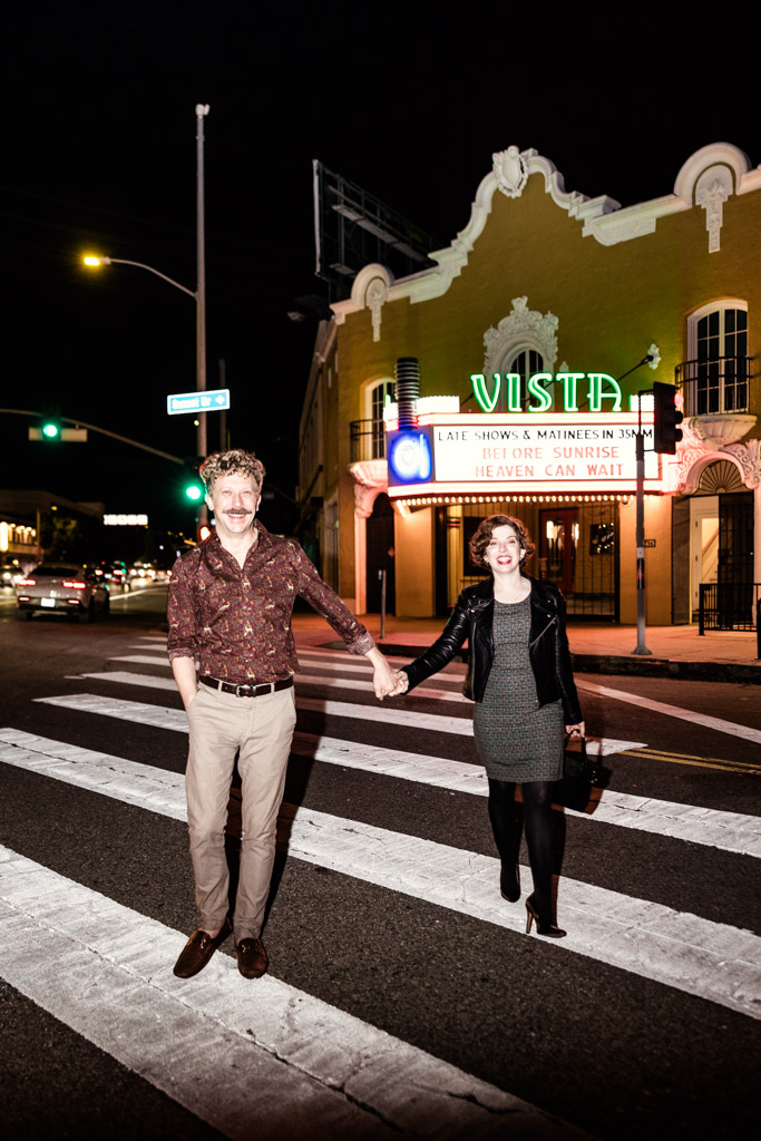 A smiling couple holds hands while crossing a street at night near the brightly lit Vista theater.