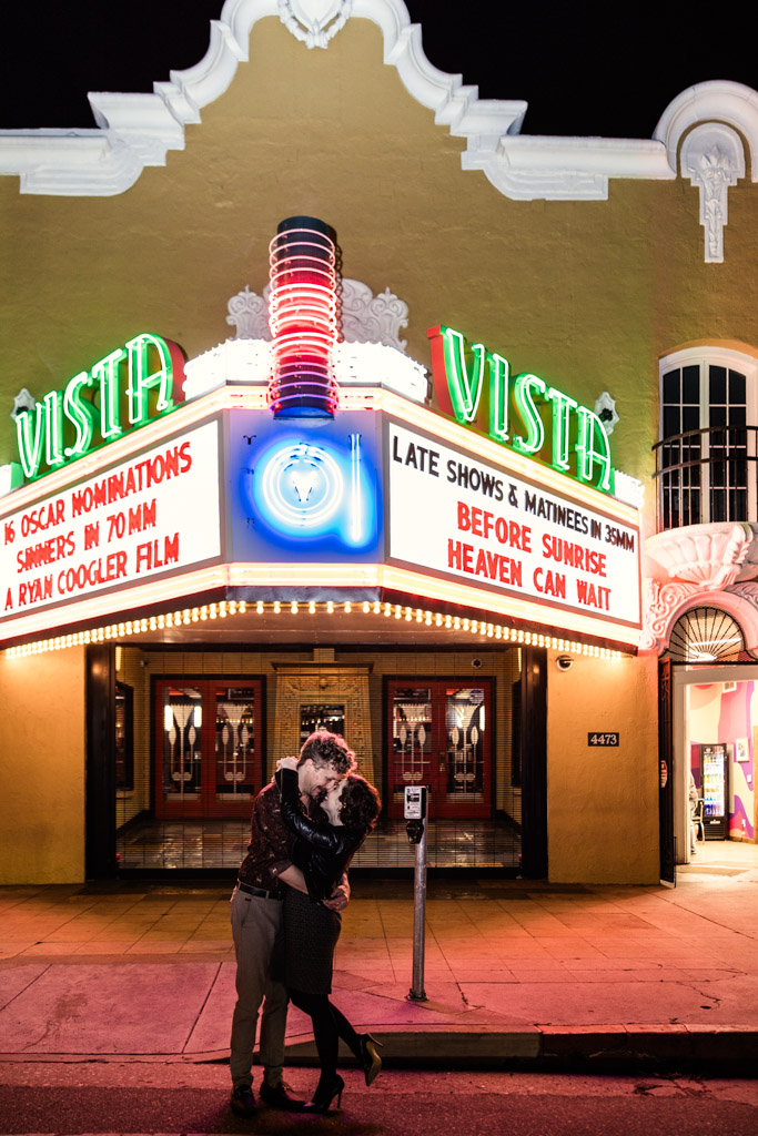Engaged couple embraces in front of the brightly lit marquee of the Vista Theatre at night