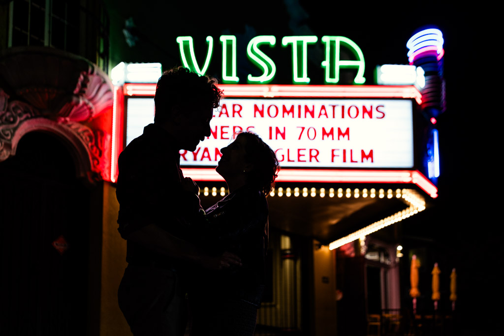 Silhouetted couple stands in front of a brightly lit Vista Theatre marquee at night