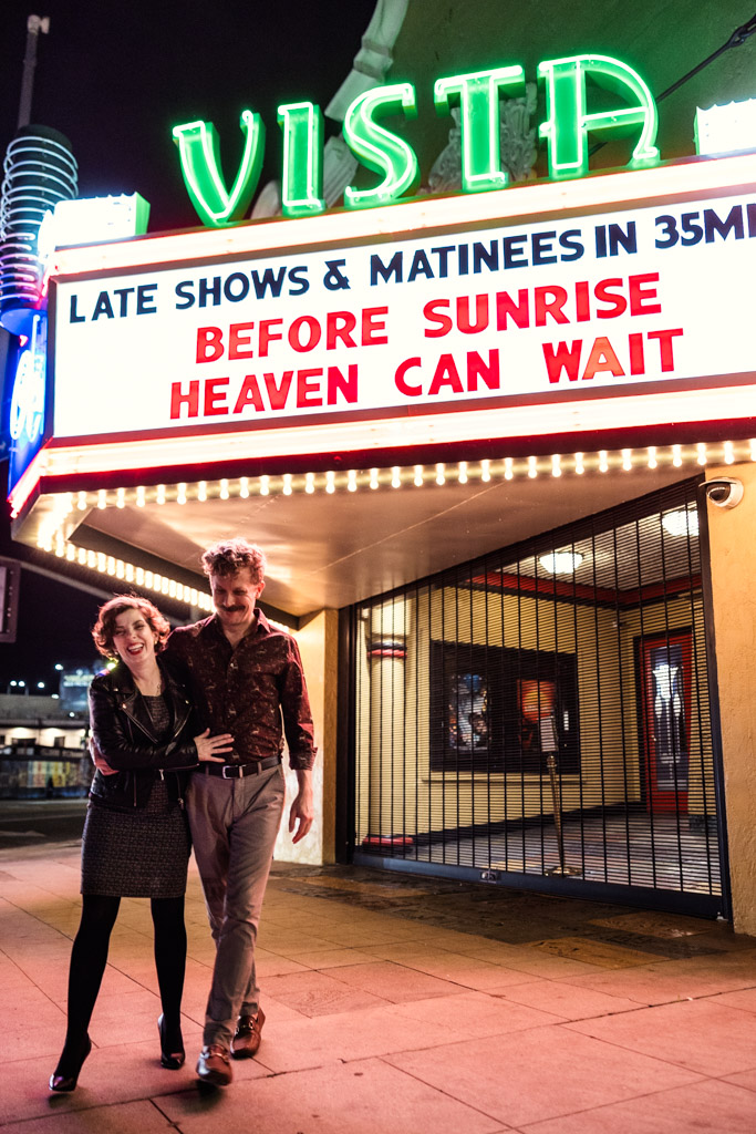 Happy engaged couple walks under the neon-lit marquee of the Vista Theater at night