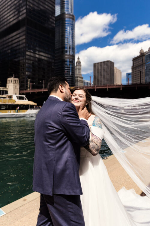 A bride and groom smile and embrace by a river with tall city buildings in the background.