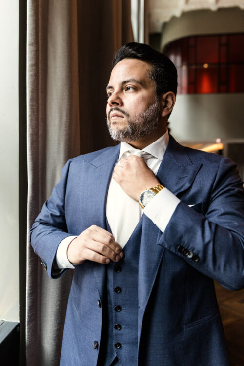 Man in a blue suit adjusting his tie while standing by a window and looking outside.