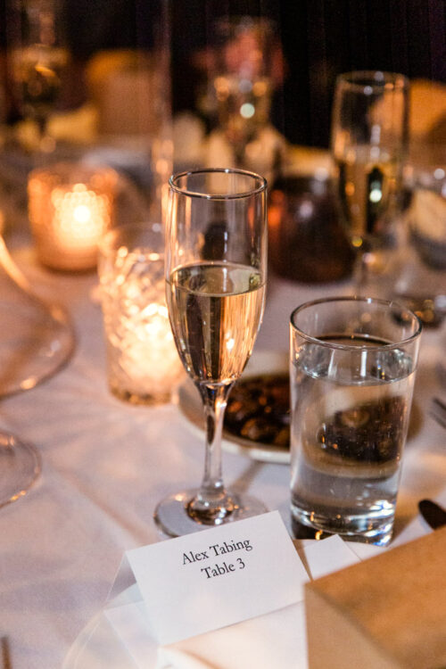 A glass of champagne and water on a candlelit table with a place card reading “Alex Tabing, Table 3.”.