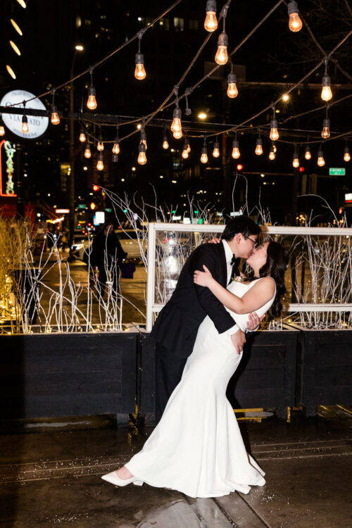 A couple in wedding attire shares a kiss under string lights on a city street at night.