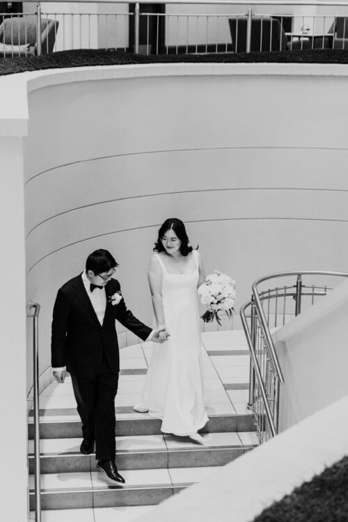 A bride and groom hold hands while walking down a staircase in a modern, indoor setting.