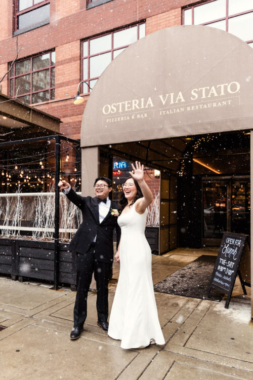 A bride and groom smile and wave under falling snow outside an Italian restaurant.