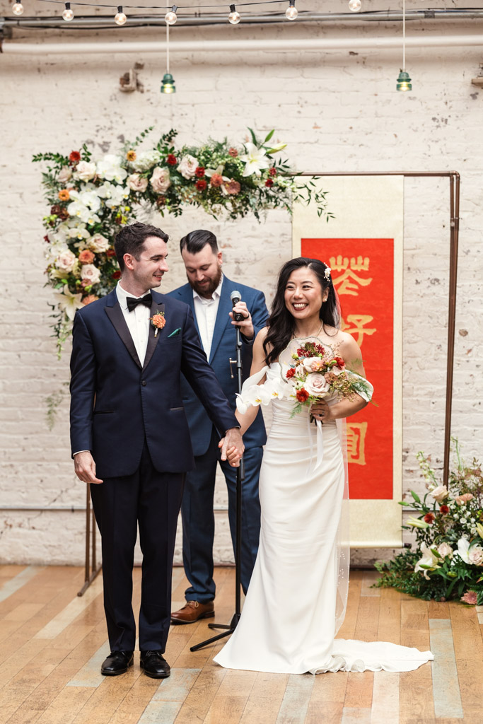 A bride and groom smile, holding hands at their wedding ceremony at The Joinery, with flowers and a red banner behind them
