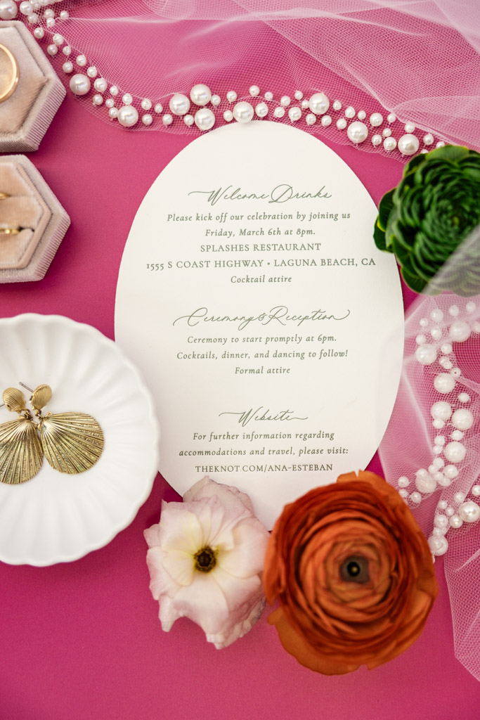 Frente al Punto wedding invitation on a pink background with flowers, jewelry, a ring box, and a pearl-trimmed veil