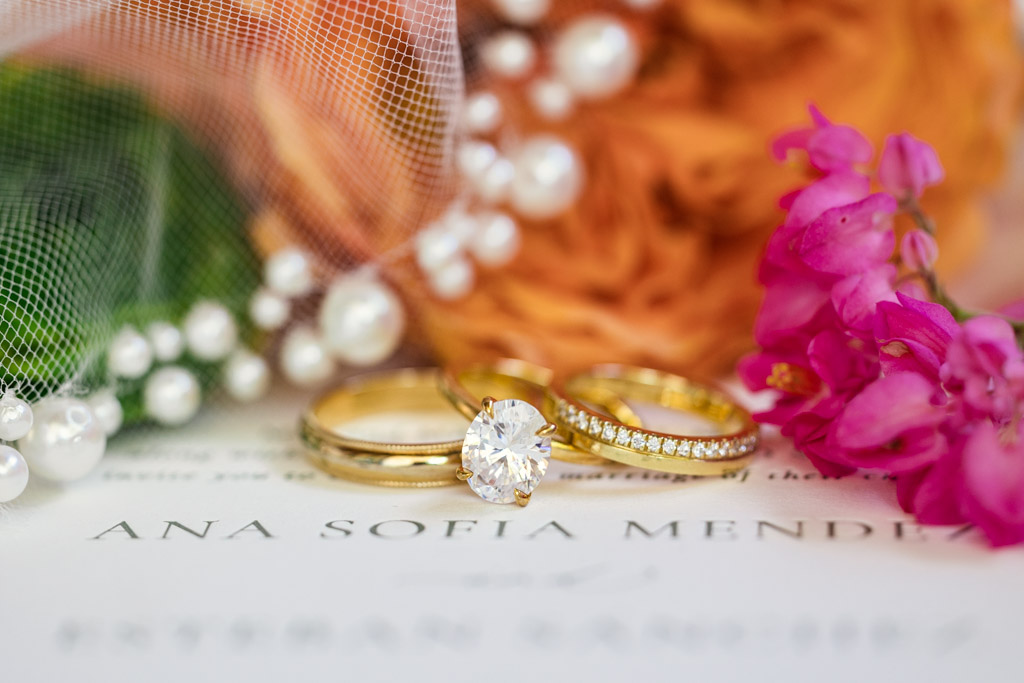 Three gold rings with diamonds rest on a wedding invitation, surrounded by flowers, pearls, and a veil for Frente al Punto wedding