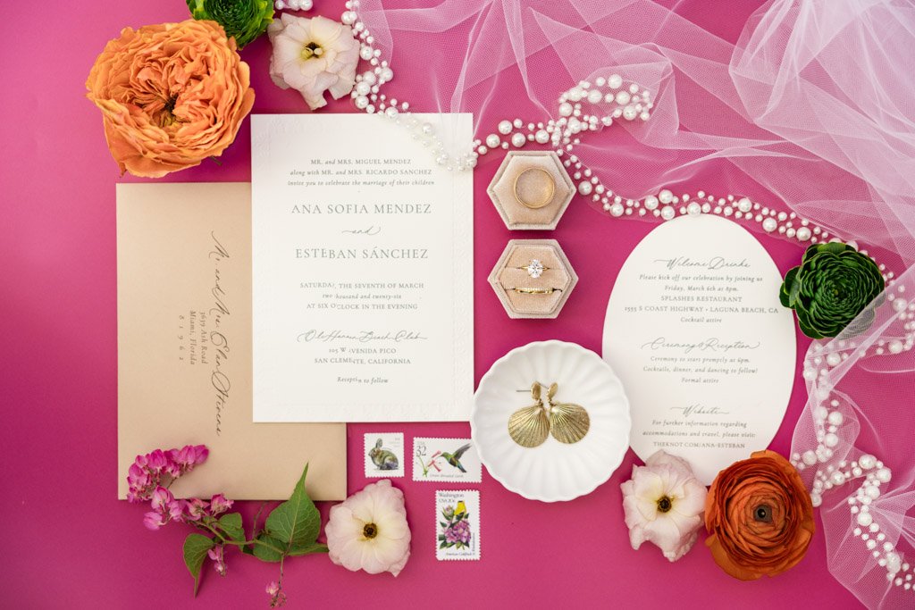 Flat lay of Frente al Punto wedding invitations, rings, stamps, flowers, and veil on a pink background