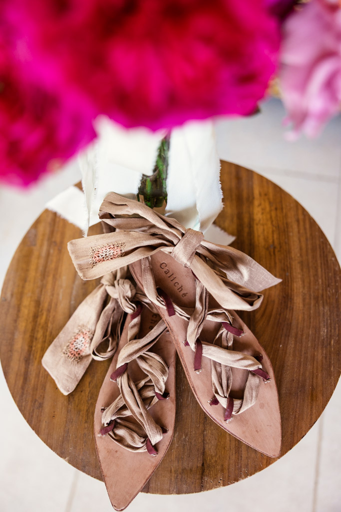 Bride's pink ribbon lace-up flats rest on a round wooden table, with bright pink flowers blurred in the foreground for a Frente al Punto wedding