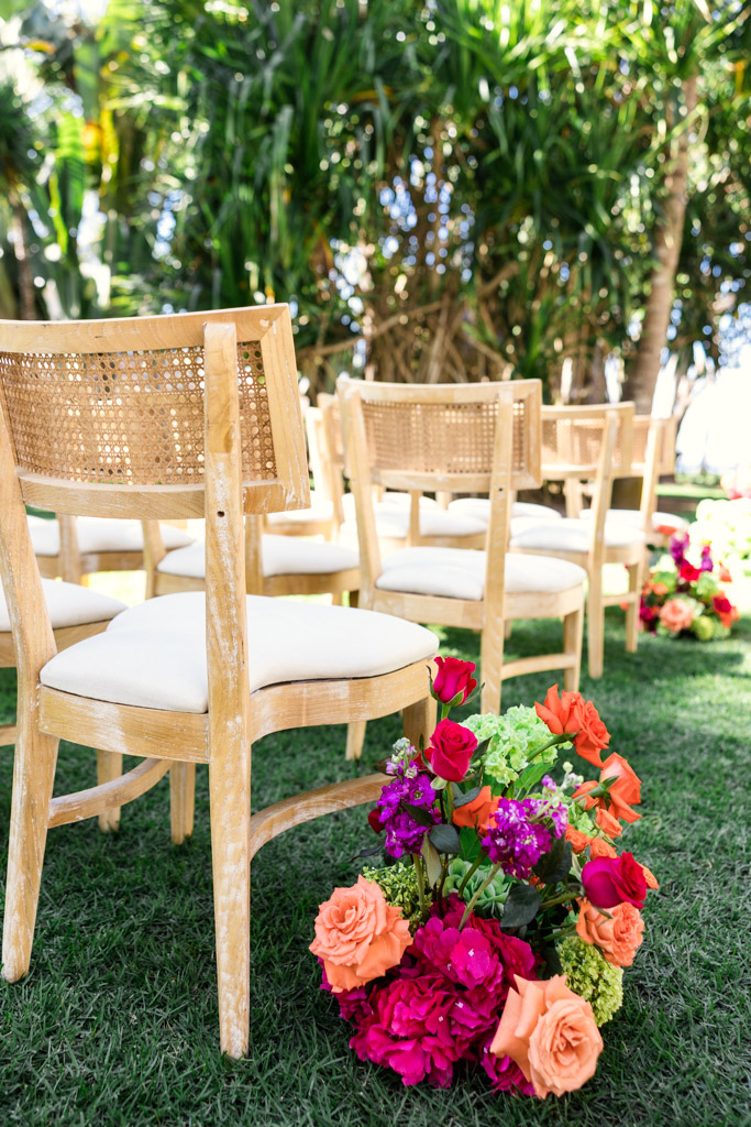 Wooden chairs arranged outdoors on grass with colorful flower arrangements beside them for a Frente al Punto wedding