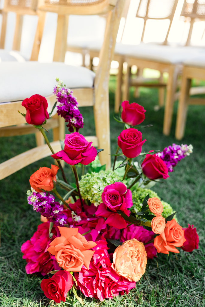 A colorful flower arrangement with red, orange, and pink blooms sits on grass by wooden chairs for a Frente al Punto wedding