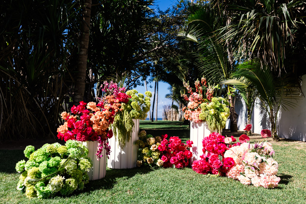 Colorful flower arrangements in white vases on green grass, surrounded by palm trees under a clear blue sky, set for a Frente al Punto wedding