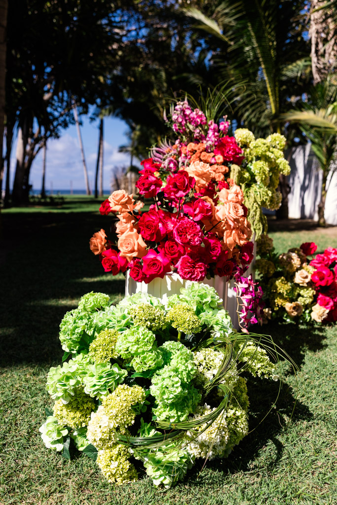Colorful flower arrangements with roses and hydrangeas displayed outdoors on grass, framed by palm trees in the background for a Frente al Punto wedding