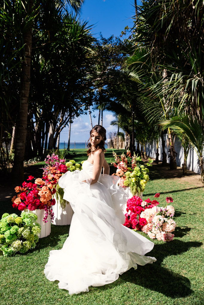 Bride in a flowing white gown poses outdoors at a Frente al Punto wedding, surrounded by colorful flower arrangements and palm trees, with the ocean in the background