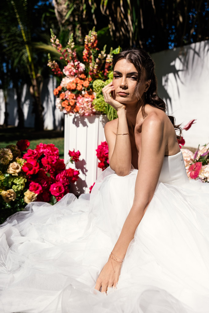 A bride in a white gown sits among colorful flowers, resting her head on her hand in bright sunlight at her Frente al Punto wedding