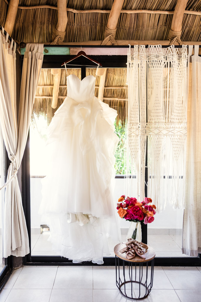 A white wedding dress hangs in a sunlit room with curtains for a Frente al Punto wedding, with bouquet of flowers rests on a small table