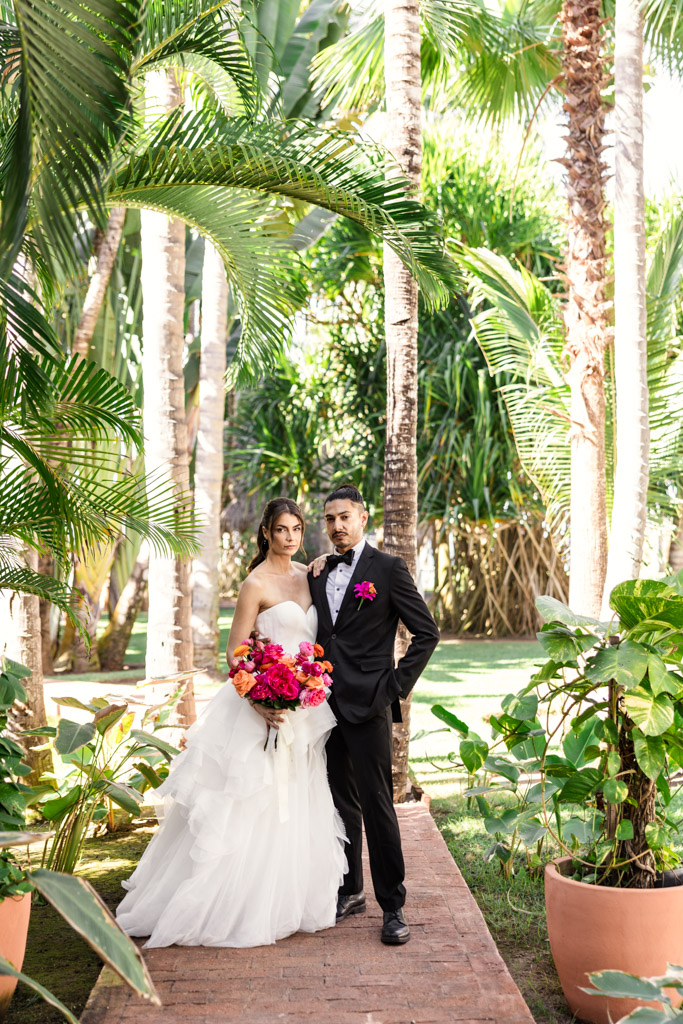 Portrait of bride and groom stand together outdoors, surrounded by tropical plants and holding a bright bouquet at their Frente al Punto wedding