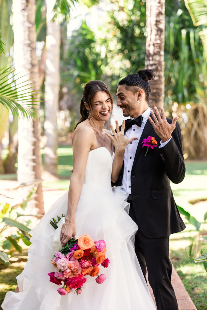 Bride and groom laughing and showing their rings outdoors at a Frente al Punto wedding, holding a colorful bouquet and surrounded by lush greenery