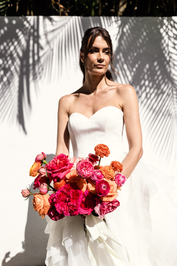 Portrait of bride in a strapless white gown holding a colorful bouquet of pink and orange flowers, standing in sunlight during her Frente al Punto wedding