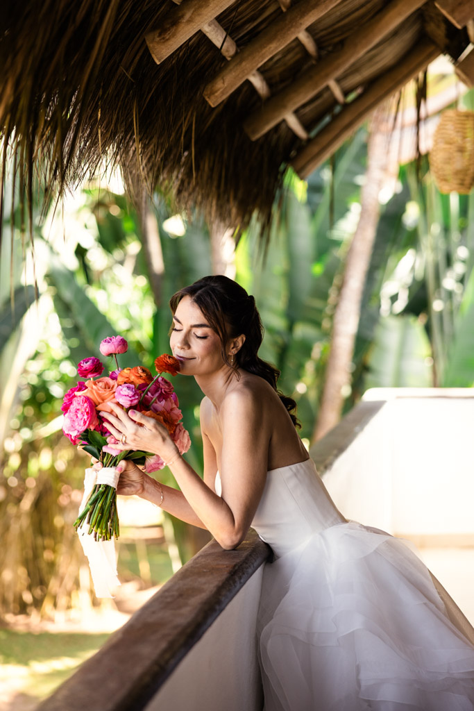 A bride in a white dress leans on a balcony during her Frente al Punto wedding, holding a colorful bouquet and smiling with eyes closed