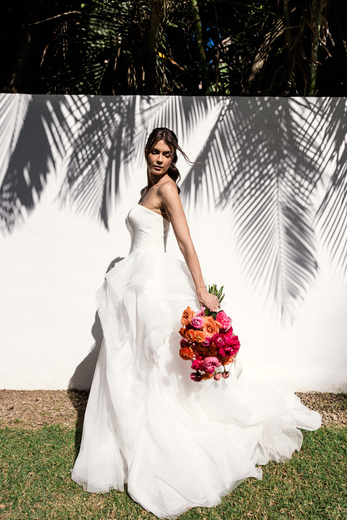 Bride in a white dress holding bright flowers, standing by a white wall with palm leaf shadows at her Frente al Punto wedding