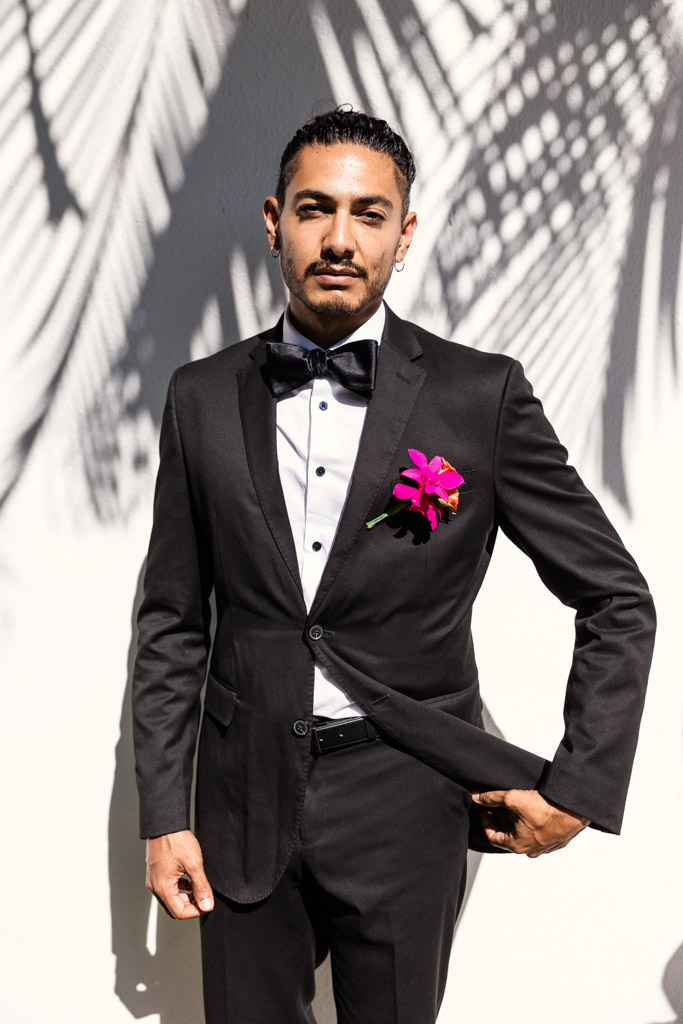 Portrait of groom in a black tuxedo with a bow tie and bright pink flower stands in sunlight with palm shadows on the wall during his Frente al Punto wedding