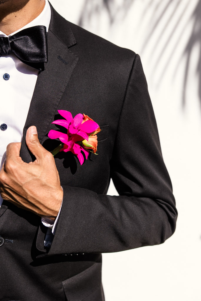 Man in a black tuxedo with a black bow tie, wearing a bright pink boutonnière, stands against a white background for a Frente al Punto wedding