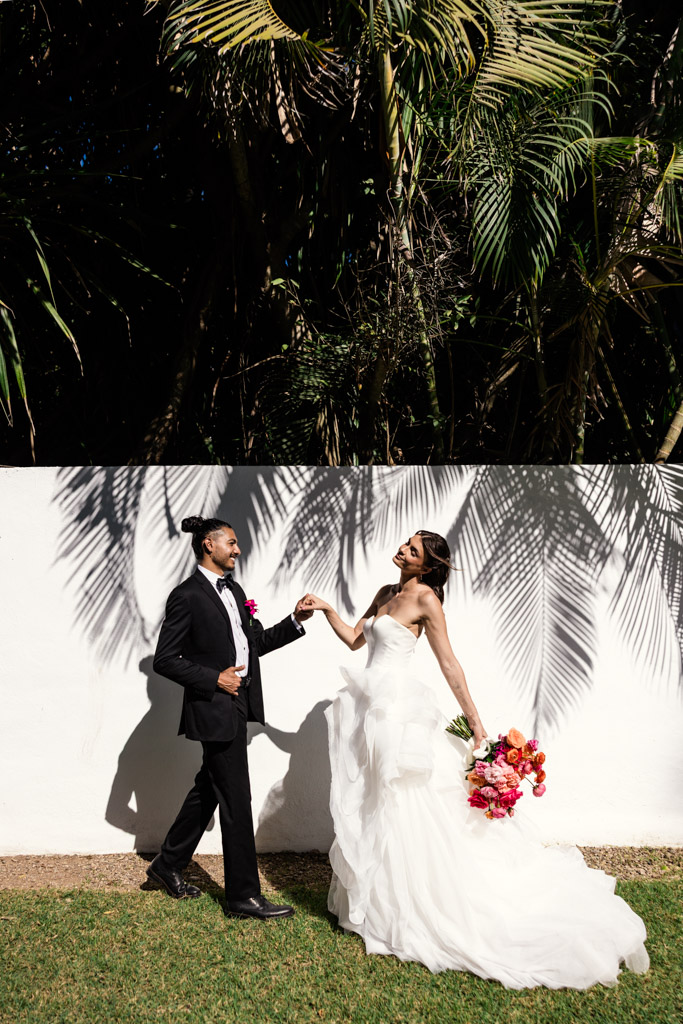 A bride and groom smile and pose together outdoors at their Frente al Punto wedding, with palm tree shadows cast on a white wall behind them