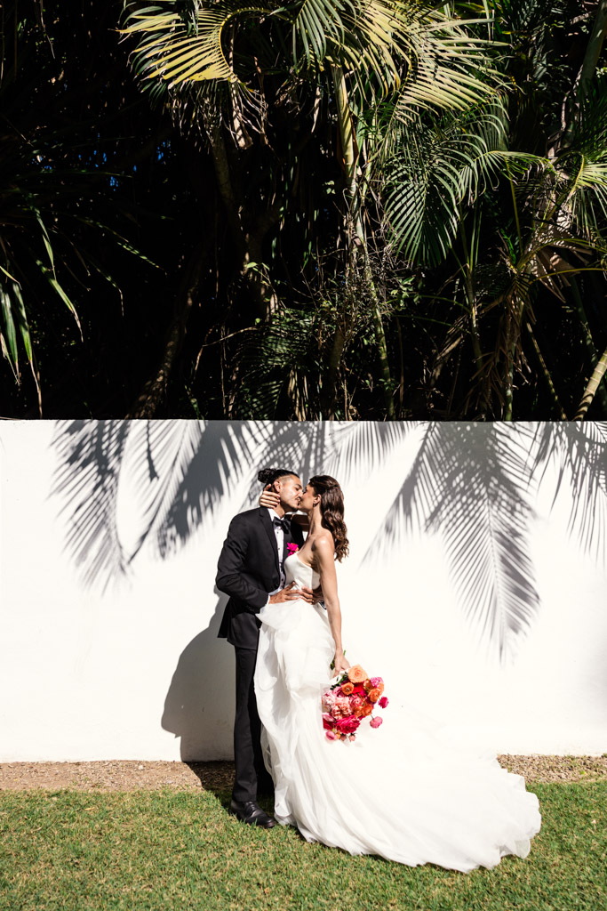 A bride and groom share a kiss outdoors, in front of a white wall with palm tree shadows during their Frente al Punto wedding