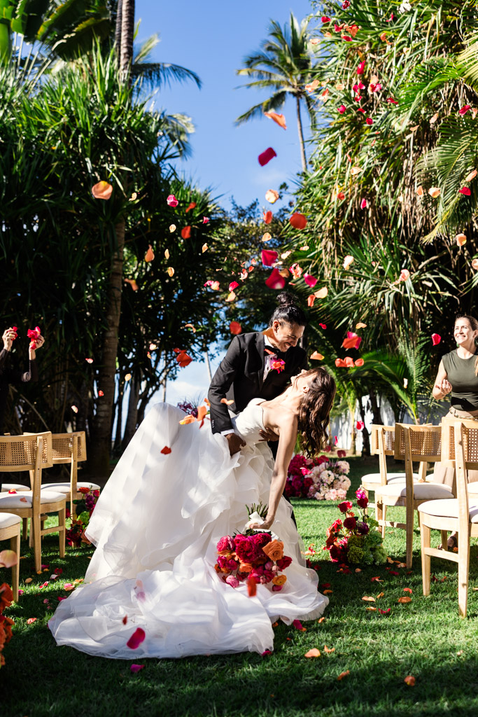 A groom dips and kisses his bride outdoors as flower petals fall around them during a joyful Frente al Punto wedding celebration