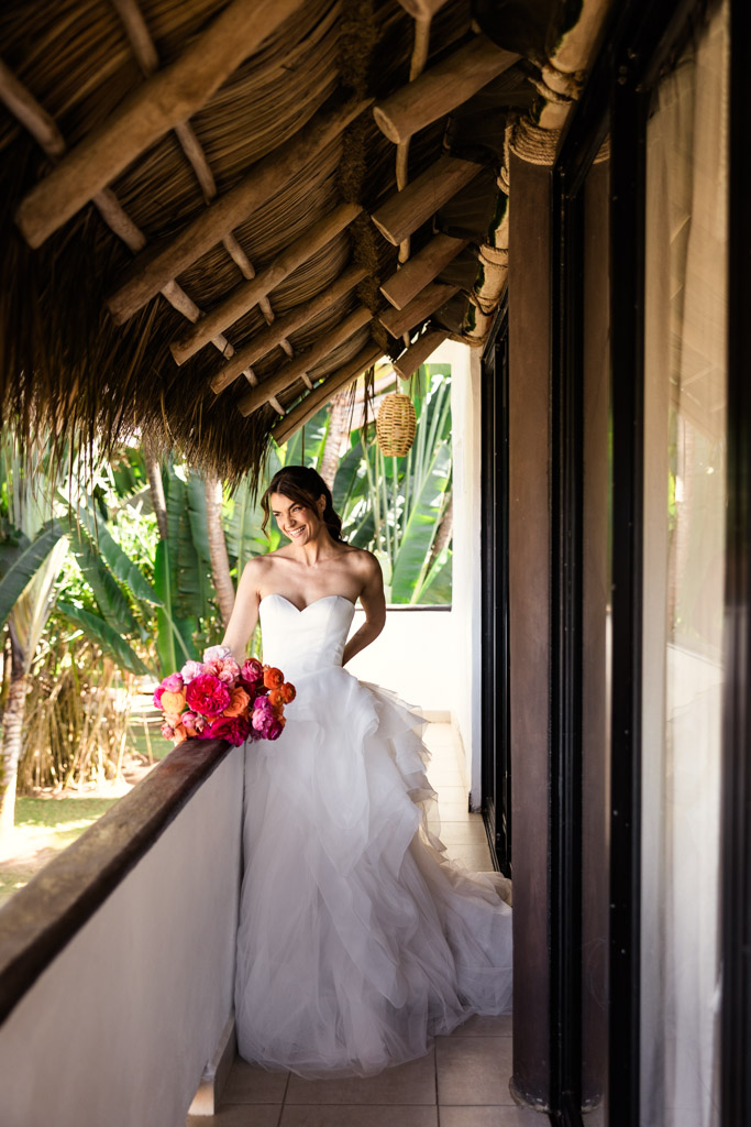Bride in a strapless white gown holding a vibrant bouquet, standing on a tropical balcony under a thatched roof during her Frente al Punto wedding