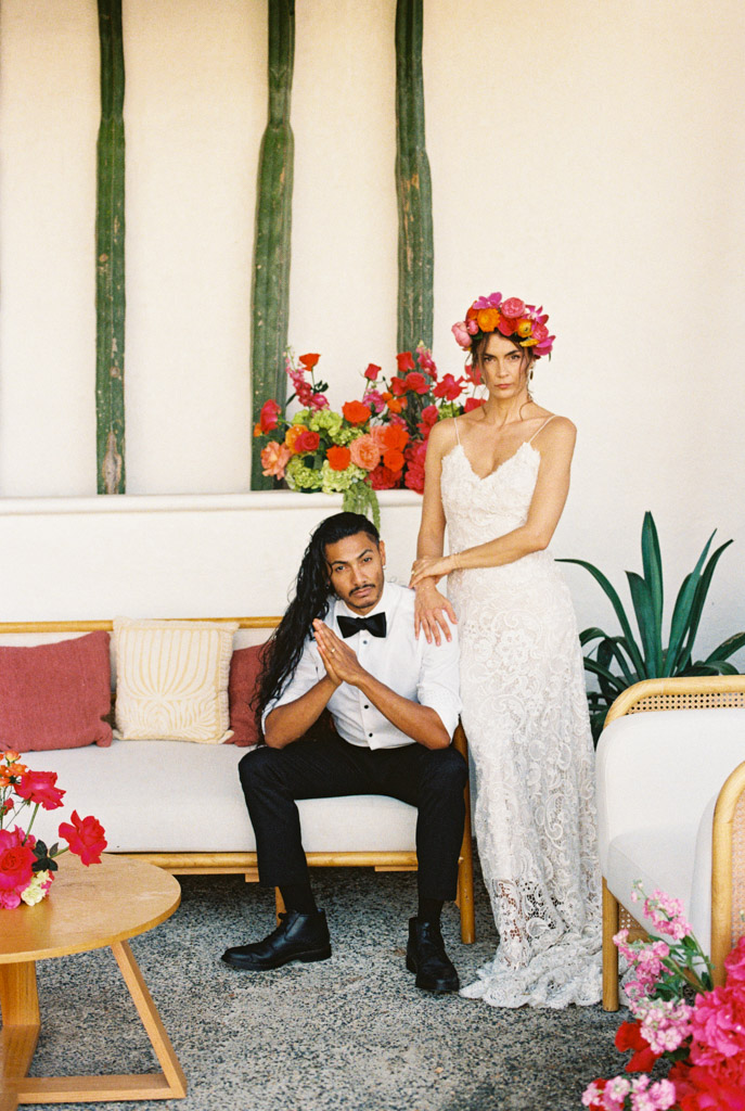 Portrait of bride in a lace dress and floral crown standing by a seated groom indoors, surrounded by bright flowers at their Frente al Punto wedding