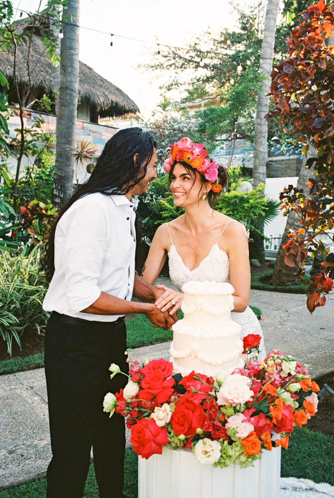 A smiling couple cuts a white wedding cake, surrounded by colorful flowers, at their Frente al Punto wedding in a lush outdoor setting