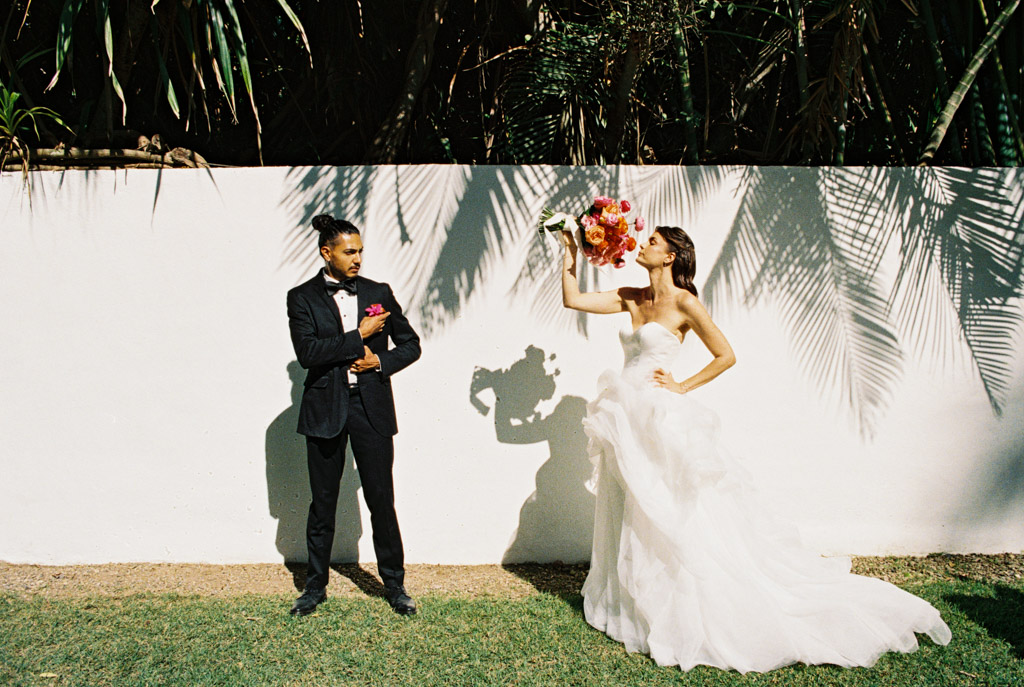 Bride in a white dress holds a bouquet, casting a shadow on a white wall, while groom in black suit stands nearby at their Frente al Punto wedding
