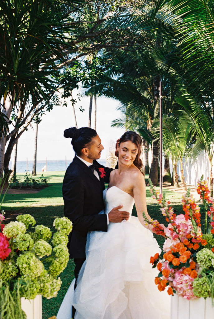 A bride and groom smile together outdoors, surrounded by lush greenery and colorful flowers at their Frente al Punto wedding
