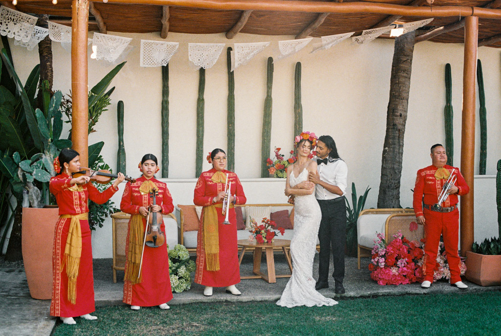 Bride and groom embrace at their Frente al Punto wedding while a mariachi band in red outfits plays music outdoors, surrounded by lush plants